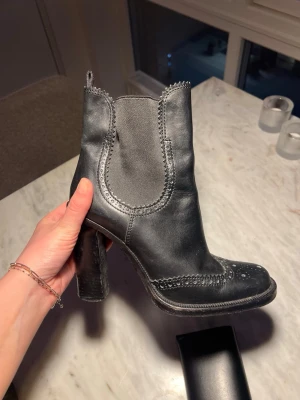 Svarta stövletter från Miu Miu - Snygga svarta stövletter från Miu Miu i äkta läder med hög klack och broguedetaljer. Resår på sidorna för enkel påtagning och en cool, chunky siluett. Perfekta för dig som vill ha en edgy och trendig look.