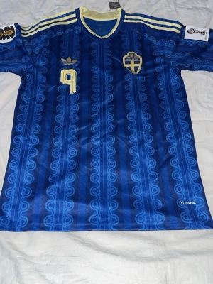 Blå Sverige fotbollströja Adidas - Säljer en blå fotbollströja från Adidas med Sveriges landslagsemblem och nummer 9 i gult. Tröjan har gul krage, gula detaljer och mönstrad design. Namnet ISAK står på ryggen. Perfekt för dig som älskar fotboll och vill stötta Sverige.