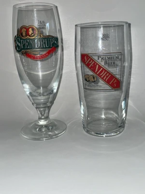Spendrups ölglas i olika modeller - 80 kr glas  ölglas från Spendrups, en med fot och en utan. Båda är transparenta med tryck i grönt, rött och guld. Glasen har Spendrups logotyp och texten 'Premium Beer' samt dekorativa detaljer. Perfekt för att samla eller använda till dryck.
