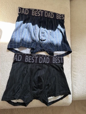 2-pack kalsonger Kappahl BEST DAD S - Två par boxerkalsonger från Kappahl i storlek S. Ena paret är mörkblå och det andra svart, båda med tunna vita ränder och bred resår med texten 'BEST DAD' i grått. Skön passform och klassisk design, perfekta för dig som vill ha något personligt och stilrent. Materialet är mjukt och stretchigt.