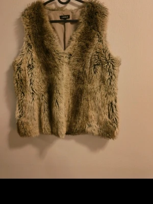 Beige fuskpälsväst från Lindex - Snygg beige fuskpälsväst från Lindex med mjukt, fluffigt material och klassisk V-ringad design. Västen har en slät, ljus innerfodring och är perfekt för att addera lite extra edge till din outfit. Passar dig som gillar trendiga och mysiga plagg.