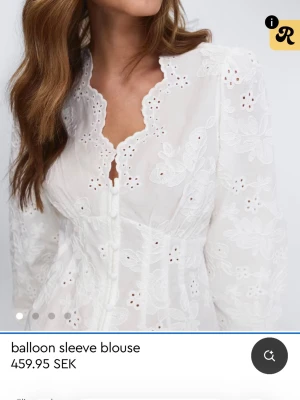 Vit broderad blus med ballongärm - Romantisk vit blus med broderade blommor och hålmönster. Blusen har ballongärmar, markerad midja och en vågig kant vid halsringningen. Knäppning framtill och lätt transparent material ger en luftig känsla.