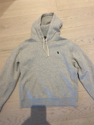  Polo Ralph Lauren Grå Hoodie - Snygg ljusgrå hoodie från Polo Ralph Lauren med klassisk marinblå logga broderad på bröstet. Hoodien har huva med vita dragsnören och en stor magficka. Tillverkad i mjuk bomullsblandning som är perfekt för chill dagar.