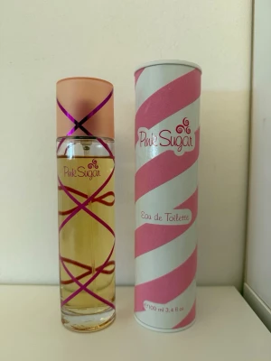 Pink Sugar Eau de Toilette 100 ml - Säljer en söt och lekfull parfym från Pink Sugar i en cylinderformad glasflaska med rosa och lila detaljer. Flaskan har ett persikofärgat lock och kommer med en matchande randig förpackning i rosa och vitt. Innehåller 100 ml Eau de Toilette.