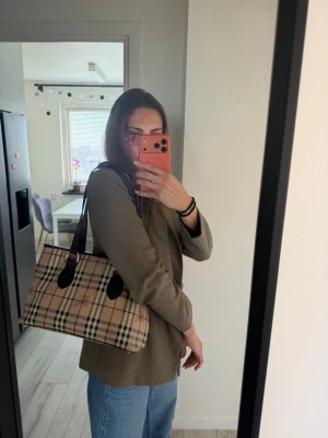 Rutig handväska från Burberry - Säljer en klassisk handväska från Burberry i beige med det ikoniska rutmönstret och svarta detaljer. Väskan har svarta handtag och är tillverkad i canvas med läderdetaljer. Perfekt storlek för att få plats med allt du behöver och ger en tidlös look.