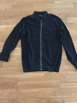 Svart zip-tröja från Riley - Stilren svart cardiganjacka från Riley med hel dragkedja framtill och klassisk krage. Tillverkad i mjukt material och har långa ärmar. Perfekt för en clean och enkel look.