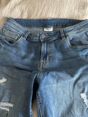 Blå slitna jeans från Kappahl - Ett par blå jeans från Kappahl med slitningar och hål på benen för en trendig och avslappnad look. Klassisk femficksmodell med dragkedja och knapp. Perfekta för dig som gillar en casual och edgy stil.