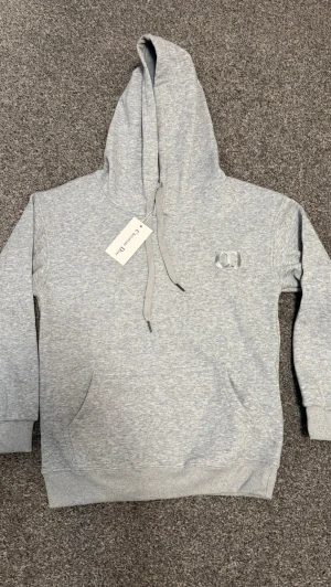 Grå hoodie - Snygg ljusgrå hoodie med broderad logga på bröstet. Klassisk passform med stor huva, dragsko och känguruficka. Tillverkad i mjuk bomullsblandning för extra komfort. Perfekt för en avslappnad och trendig look.