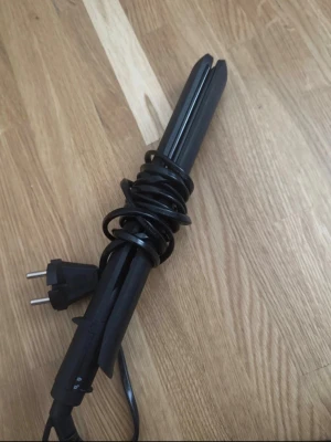 Svart plattång för hårstyling - En svart plattång med sladd och europeisk kontakt, perfekt för att styla håret. Plattången har en smal och avlång form med två plattor som möts när man klämmer ihop den. Tillverkad i plast och metall.