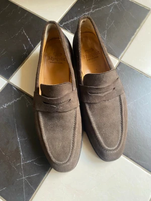 Bruna mockaloafers från Myrqvist - Snygga bruna loafers i mocka från Myrqvist med klassisk penny strap och stilren design. Skorna har en mjuk innersula i ljust läder och en elegant, tidlös form som passar till många olika outfits. Perfekta för dig som gillar stilrena och bekväma skor. De är i mycket bra skick.