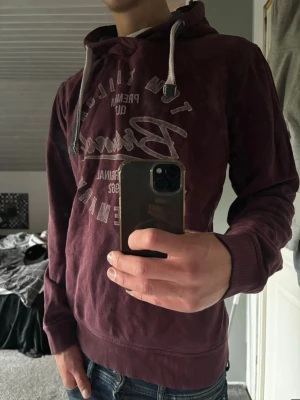 Tom Tailor hoodie - Säljer en vinröd hoodie från Tom Tailor med tryck på bröstet och vita dragsnören i huvan. Hoodien har ribbade muddar vid ärmslut och nederkant samt en klassisk känguruficka framtill. Perfekt för dig som gillar en avslappnad och snygg stil.