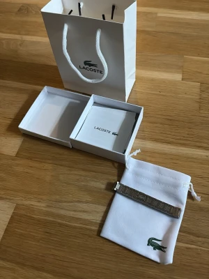 Silverfärgat armband från Lacoste - Snyggt silverfärgat armband från Lacoste med stilren design och diskret logotyp. Armbandet är i metall och kommer med originalpåse, ask och presentpåse från Lacoste. Perfekt accessoar för dig som gillar klassisk och modern stil.