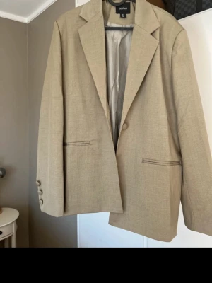 Beige kavaj från Boohoo - Stilren beige kavaj från Boohoo med klassisk passform, enkelknäppning och två fickor framtill. Kavajen har tre matchande knappar vid ärmslutet och är helfodrad. Perfekt för en trendig och clean look.