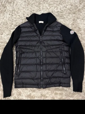 Svart dunväst från Moncler - Snygg svart dunväst från Moncler med stickade svarta ärmar och hög krage. Västen har två dragkedjeförsedda fickor framtill och ett Moncler-märke på ena ärmen. Perfekt för lager-på-lager och riktigt trendig look.