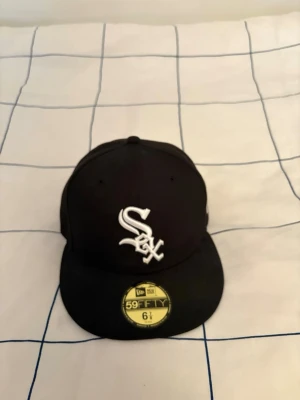 Svart New Era White Sox keps - Säljer en svart New Era 59FIFTY keps med White Sox-logga broderad i vitt framtill. Klassisk flat peak och strukturerad passform. Kepsen är tillverkad i bomull och polyester, perfekt för dig som gillar sportig streetstyle.