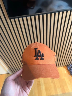 Orange LA-keps från New Era - Snygg orange keps från New Era med LA-logga broderad i svart framtill. Klassisk böjd skärm och justerbar rem bak. Tillverkad i bomullstyg som ger en avslappnad och sportig vibe. Perfekt accessoar för dig som gillar streetwear och vill sticka ut. Pris kan diskuteras:)