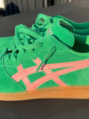Gröna Asics Skyhand sneakers, storlek 44 - Snygga gröna Asics Skyhand sneakers i mocka med rosa detaljer och klassiska Asics-stripes på sidan. Skorna har en låg siluett, gröna skosnören och en gummisula i naturfärg. Perfekta för dig som vill sticka ut med färg och sportig vibe. Skorna är sparsamt använda. 