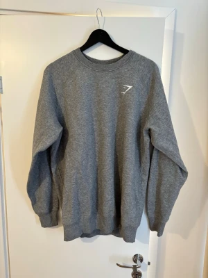 Grå sweatshirt från Gymshark - En stilren grå sweatshirt från Gymshark med rund halsringning och diskret logga på bröstet. Tröjan har ribbade muddar vid ärmslut och nederkant, perfekt för en avslappnad och sportig look. Tillverkad i mjukt material för extra komfort.