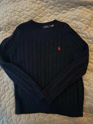 Mörkblå kabelstickad tröja Polo Ralph Lauren - Mörkblå kabelstickad tröja från Polo Ralph Lauren med rund halsringning och klassisk röd broderad logga på bröstet. Tröjan har långa ärmar och ribbade muddar vid ärmslut och nederkant. Perfekt för dig som gillar tidlös och stilren design.