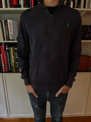 Mörkblå halvzip från Ralph Lauren - Mörkblå tröja med halv dragkedja och hög krage från Ralph Lauren. Klassisk grön logga broderad på bröstet och ribbade muddar vid ärmslut och nederkant. Perfekt till jeans för en clean och stilren look.