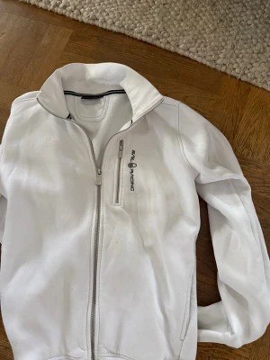 Vit Hoodie från Sail Racing - Vit hoodie från Sail Racing med hög krage och dragkedja framtill. Hoodien har en bröstficka med dragkedja och logga på vänster sida. Tillverkad i ett mjukt syntetmaterial och har långärmad design.