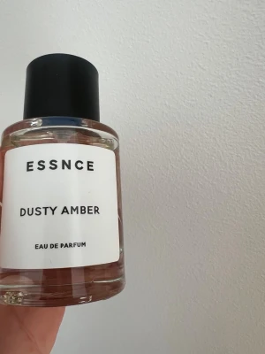 ESSNCE Dusty Amber Eau de Parfum - ESSNCE Dusty Amber Eau de Parfum i en stilren, rund glasflaska med svart lock och vit etikett. Doften har en elegant och minimalistisk design som passar perfekt på parfymhyllan. Flaskan är genomskinlig och visar den ljusa vätskan inuti.