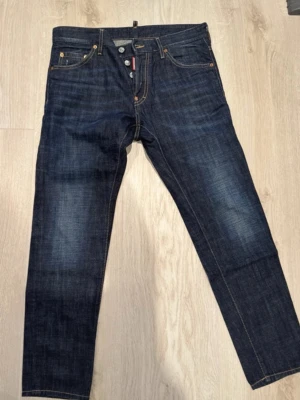 Mörkblå jeans från Dsquared2 - Säljer ett par snygga mörkblå jeans från Dsquared2 med klassisk femficksmodell och raka ben. Jeansen har tydliga kontrastsömmar och slitningar på låren för en cool look. Tillverkade i kraftigt denimtyg som ger en schysst passform. Sparsamt använda. Är 183 cm lång och bär storlek S/M. 