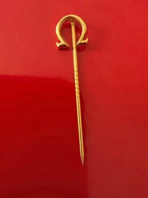 Guldfärgad Omega slipsnål - Snygg guldfärgad slipsnål med Omega-loggan i toppen. Tillverkad i metall med elegant och stilren design. Perfekt accessoar för dig som gillar klassiska detaljer och vill lyfta din outfit med något unikt. Är från ett OS på 80-talet, då min pappa fått den, har stämplar på baksidan jag inte kan utläsa.