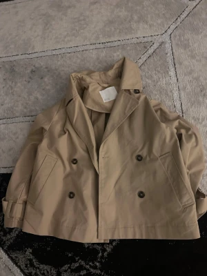 Kort beige trenchcoat med dubbelknäppning - Stilren kort trenchcoat i beige med klassisk dubbelknäppning och bred krage. Jackan har två sidofickor och justerbara ärmslut. Perfekt för en trendig och avslappnad look. 