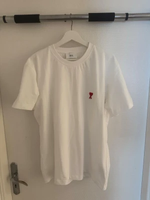 Vit t-shirt från Ami Paris med rött hjärta - Klassisk vit t-shirt från Ami Paris med ett litet rött hjärta broderat på bröstet. T-shirten har rund halsringning och korta ärmar. Tillverkad i mjuk bomull för en skön och avslappnad känsla.  Storlek L men passar Storlek M också. 
