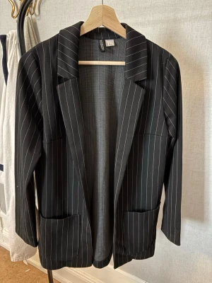 Svart kavaj med vita ränder från Gina Tricot - Snygg svart kavaj från H&M med tunna vita vertikala ränder. Klassisk modell med slag, två framfickor och rak passform. Perfekt för en stilren och trendig look. Materialet känns mjukt och följsamt.
