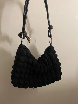 Gullig handväska  - Trendig svart handväska med puffigt, strukturerat tyg och justerbart axelband med knutdetaljer och guldfärgade metalldelar. Väskan har dragkedja och ljus insida. Perfekt för dig som gillar unika accessoarer och vill sticka ut.