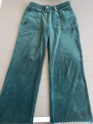 Gröna mjukisbyxor från Juicy Couture - Mysiga gröna mjukisbyxor från Juicy Couture i velour med breda ben, stora fickor framtill och dragsko i midjan. Klassisk JC-logga broderad på ena fickan. Perfekta för chill dagar och riktigt sköna att ha på sig.