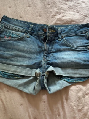 Blå denimshorts med mönstrad detalj - Säljer ett par blå denimshorts med uppvikta benslut och coola mönstrade detaljer i fickorna och baktill. Klassisk femficksmodell med knapp och dragkedja. Perfekta för varma dagar och enkel att matcha med allt.