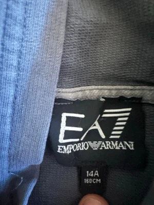 Grå hoodie från Emporio Armani EA7 - Snygg grå hoodie från Emporio Armani EA7 med klassisk huva och stor magficka. Diskret logga på bröstet och mjukt material som känns skönt mot huden. Perfekt för dig som gillar stilrena och bekväma plagg.