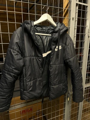 Svart dunjacka från Nike - Snygg svart dunjacka från Nike med huva och dragkedja framtill. Jackan har en quiltad design och Nike-logga på bröstet. Perfekt för kalla dagar och har en sportig look med justerbar huva och praktiska fickor.
