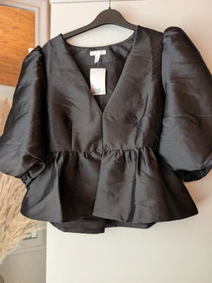 Svart puffärmad blus från H&M - Stilren svart blus från H&M med voluminösa puffärmar och v-ringning. Blusen har en markerad midja med rynk och är tillverkad i ett glansigt, satinliknande material som ger en lyxig känsla. Perfekt för dig som vill sticka ut med en trendig siluett.