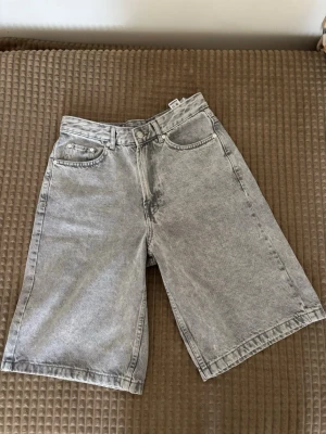 Gråa loose denimshorts - Säljer ett par gråa loose-fit denimshorts med fem fickor och bälteshällor. De har en klassisk knapp- och dragkedjestängning och är tillverkade i ett mjukt bomullsmaterial. Perfekta för dig som gillar en avslappnad och trendig stil.