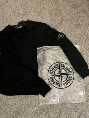 Stone Island tröja - Säljer en svart sweatshirt från Stone Island med den ikoniska kompasspatchen på vänster ärm. Tröjan har rund halsringning, ribbade muddar och är tillverkad i mjukt material. Perfekt för dig som gillar streetwear och stilrena plagg.
