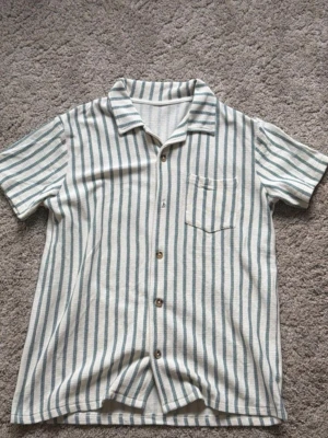 Ralph Lauren randig T-shirt. - Snygg kortärmad skjorta från Ralph Lauren med vertikala gröna och vita ränder. Skjortan har knäppning framtill med träfärgade knappar och en bröstficka. Tillverkad i ett mjukt och lätt tyg, perfekt för varma dagar. Avslappnad passform och klassisk krage.