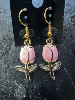 Rosa blomörhängen med strassdetaljer - Snygga örhängen i form av rosa blommor med guldfärgade detaljer och små glittrande stenar i bladen. Hängande modell med krok, perfekt för att ge din look en söt och trendig touch. Materialet är metall och emalj med strass.