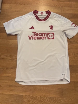 Vit Manchester United tröja från säsongen 23/24 - Snygg vit Manchester United matchtröja från Adidas med röda detaljer och logga. Tröjan har korta ärmar, rund hals med röd kant och TeamViewer-tryck på bröstet. Tillverkad i lätt och ventilerande material, perfekt för fotboll eller träning och även för samling 