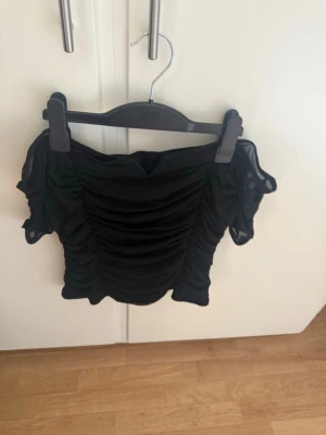Svart offshoulder  topp - Säljer en svart offshoulder topp med rynkade detaljer framtill och på ärmarna. Toppen har korta,  ärmar i mesh-liknande material som ger en trendig look. Perfekt för dig som vill sticka ut lite extra.