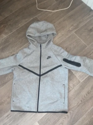 Grå Nike hoodie med dragkedja XL - Snygg grå hoodie från Nike i storlek XL med hel dragkedja framtill, svarta detaljer och fickor. Hoodien har en huva och en extra ficka med dragkedja på ena ärmen. Perfekt för en sportig och avslappnad stil.