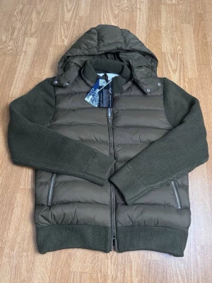 Olivgrön pufferjacka med stickade ärmar - Snygg olivgrön pufferjacka med avtagbar huva och stickade ärmar samt muddar. Jackan har två dragkedjeförsedda fickor och quiltad kropp i glansigt material. Perfekt för kalla dagar och ger en modern look.