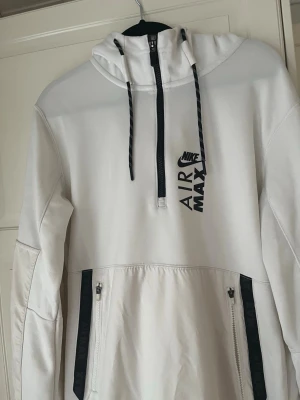 Vit Nike Air Max hoodie med dragkedja - Snygg vit hoodie från Nike Air Max med svart logga på bröstet. Hoodien har huva med dragsko, halv dragkedja framtill och två svarta dragkedjefickor. Perfekt för en sportig och avslappnad stil.