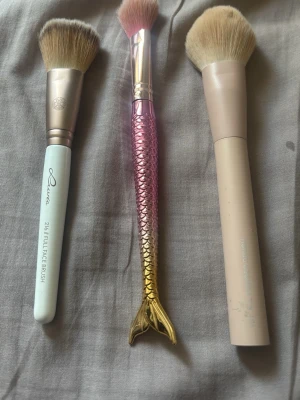 Set med tre sminkborstar - Säljer ett set med tre sminkborstar i olika stilar. En borste har ljusblått handtag och täta borst, en har ett metallicfärgat handtag i form av en sjöjungfrustjärt i rosa och guld, och en har ett beige handtag. Alla har mjuka, täta borst i beige eller rosa toner. Perfekt för applicering av puder, rouge eller bronzer.