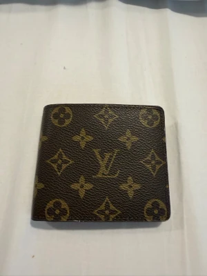  brun monogram plånbok - Säljer en klassisk brun plånbok med det ikoniska monogrammönstret i guld. Plånboken är i läder och har flera fack för kort och sedlar. Insidan är ljusbrun. Perfekt accessoar för dig som gillar lyxiga detaljer.