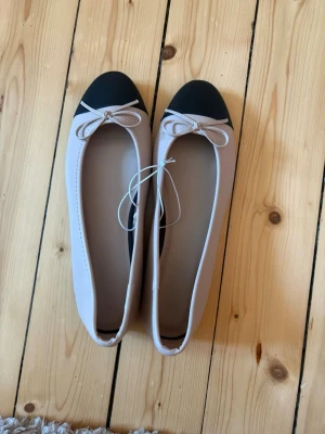 Beige ballerinaskor med svarta tår - Snygga ballerinaskor i beige med svarta tår och dekorativa rosetter framtill. Skorna har en klassisk rundad form och är tillverkade i ett slätt syntetmaterial. Perfekta för en stilren och enkel look.