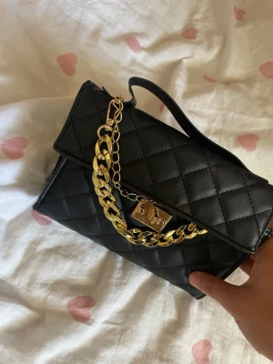 Svart quiltad handväska med guldkedja - Snygg svart handväska i quiltat mönster med en chunky guldfärgad kedja och metallspänne framtill. Väskan har ett elegant och stilrent utseende med ett kort handtag upptill. Perfekt accessoar för att lyfta din outfit. Dock kan man inte stänga väskan, hållaren till den är sönder, men kan man fylla på väskan annars som vanligt ändå. Därför säljer jag den billigt:)
